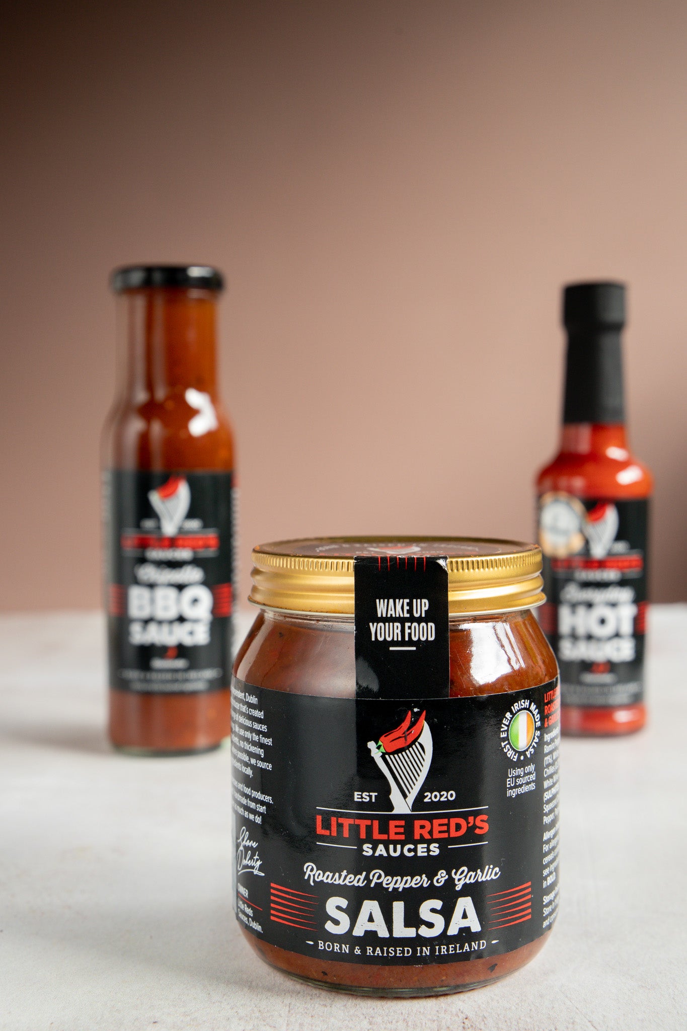 Focused close-up of Little Red's Gluten-Free Slow Roasted Pepper & Garlic Salsa, with other sauces softly blurred in the background. The salsa's label is prominent, showcasing its key features of slow-roasted peppers and garlic, underlining its gluten-free attribute, and appealing to health-conscious and flavor-seeking consumers
